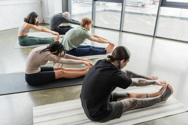 Vue latérale de personnes interraciales faisant assis vers l'avant plier asana sur des nattes en cours de yoga — Photo de stock
