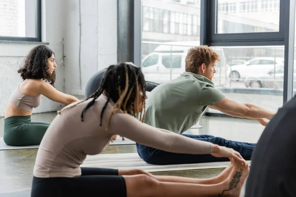 Visão lateral de jovens interracial alongamento enquanto fazendo sentado para a frente curva asana no estúdio de ioga — Fotografia de Stock