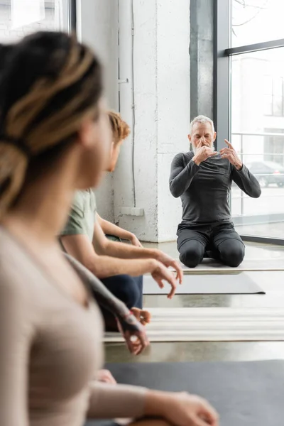 Mature coach practicing nostril breathing near blurred interracial group in yoga class — Stock Photo