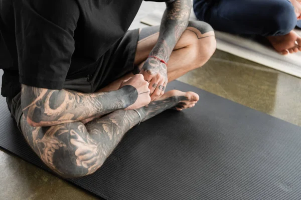 Vista ritagliata di uomo tatuato seduto nel fuoco log asana sul tappeto in classe di yoga — Foto stock