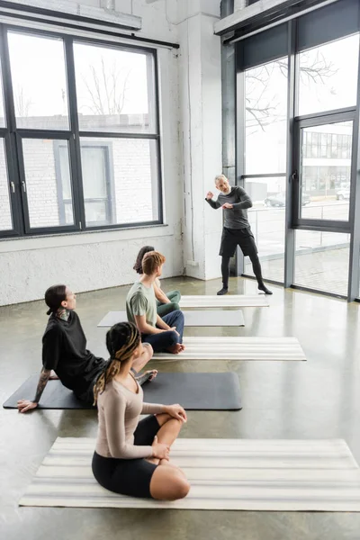 Entrenador maduro hablando con grupo interracial borroso en clase de yoga - foto de stock