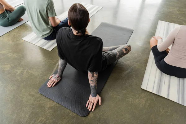 Вид сверху на татуированного человека, сидящего на коврике рядом с межрасовыми людьми в классе йоги — стоковое фото