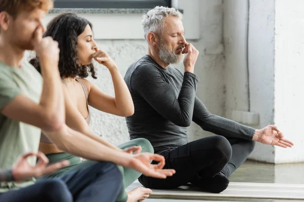 Uomo maturo che pratica la respirazione narice e mudra gyan vicino a persone interrazziale in studio di yoga — Foto stock