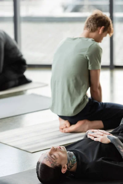 Татуйований чоловік лежить на килимку біля розмитих людей в класі йоги — стокове фото