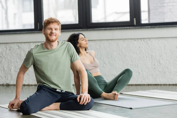 Uomo rosso sorridente che guarda la macchina fotografica vicino alla donna mediorientale in studio di yoga — Foto stock