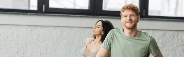 Позитивный рыжий мужчина смотрит в камеру рядом с женщиной с Ближнего Востока в студии йоги, баннер — стоковое фото
