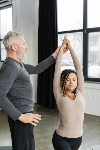 Allenatore di mezza età in piedi vicino alla donna afro-americana che fa Crescent Lunge asana in classe di yoga — Foto stock