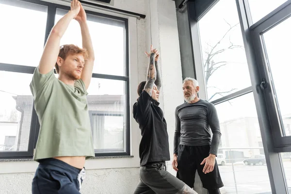 Middle aged coach talking to young men doing Crescent Lunge asana in yoga studio — Stock Photo