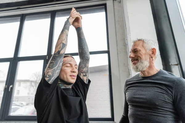 Улыбающийся тренер средних лет, смотрящий на татуированного мужчину, делающего полумесяц выпад асану в классе йоги — стоковое фото
