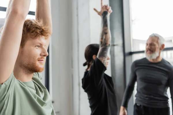 Smiling redhead man standing in Crescent Lunge asana near blurred group in yoga studio — Stock Photo