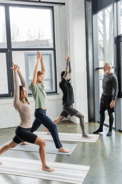 Interrassische Menschen stehen in Crescent Lunge Asana in der Nähe eines reifen Trainers im Yoga-Kurs — Stockfoto