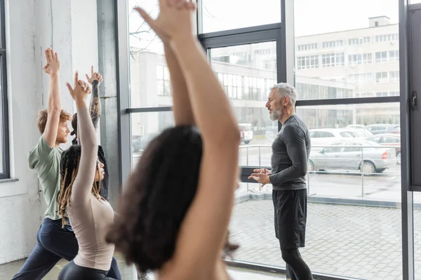 Vue latérale du coach mature parlant à des personnes multiethniques debout dans la pose de Crescent Lunge dans un studio de yoga — Photo de stock