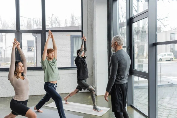 Trainer mittleren Alters, der in der Nähe einer multiethnischen Gruppe steht und Crescent Lunge übt, posiert im Yogastudio — Stockfoto