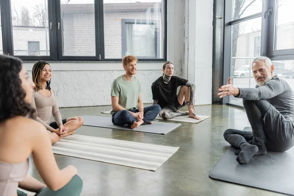 Smiling coach pointing at blurred middle eastern woman near people in yoga class — Stock Photo