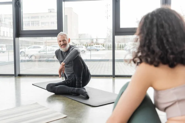 Alegre entrenador maduro mirando a la mujer borrosa mientras está sentado en la estera en el estudio de yoga - foto de stock
