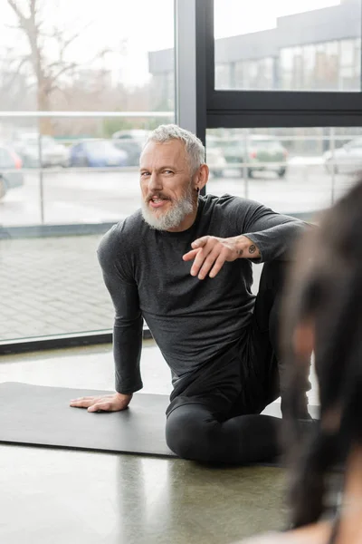 Smiling mature coach pointing at blurred african american woman in yoga class — Stock Photo