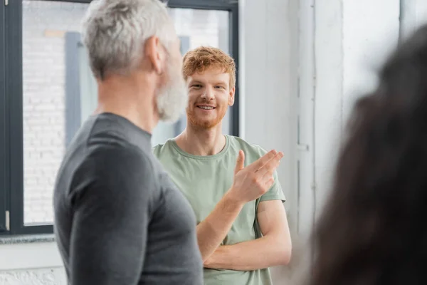 Улыбающийся рыжий мужчина разговаривает с размытым тренером в классе йоги — стоковое фото
