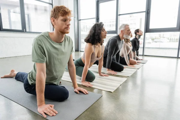 Rousse homme pratiquant Half Pigeon asana près de personnes multiethniques en classe de yoga — Photo de stock