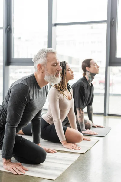 Hombre de mediana edad practicando Half Pigeon asana cerca de personas multiétnicas en clase de yoga - foto de stock