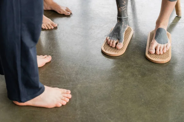Cropped view of tattooed man standing with bare feet on sadhu nail board near faceless people in yoga studio — Stock Photo