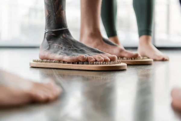 Частковий вигляд людини з татуюванням на ногах, що стоять на дошці для нігтів біля людей в студії йоги — стокове фото