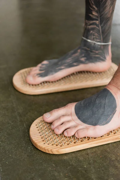 Vista recortada del hombre descalzo con las piernas tatuadas de pie en el tablero de clavos sadhu - foto de stock