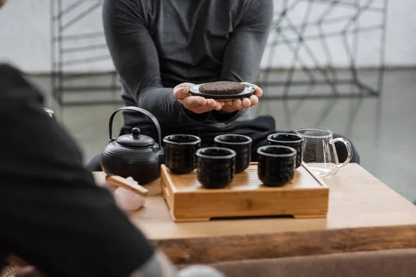 Cropped view of man holding compressed puer tea near traditional teapot and cups — Stock Photo