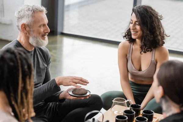 Tatuado homem com barba cinza segurando comprimido puer chá perto alegre médio oriental mulher no estúdio de ioga — Fotografia de Stock