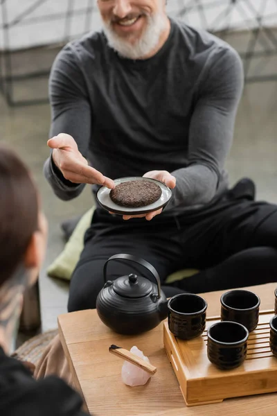 Vista recortada de hombre feliz de mediana edad con barba gris sosteniendo té puer comprimido en estudio de yoga - foto de stock
