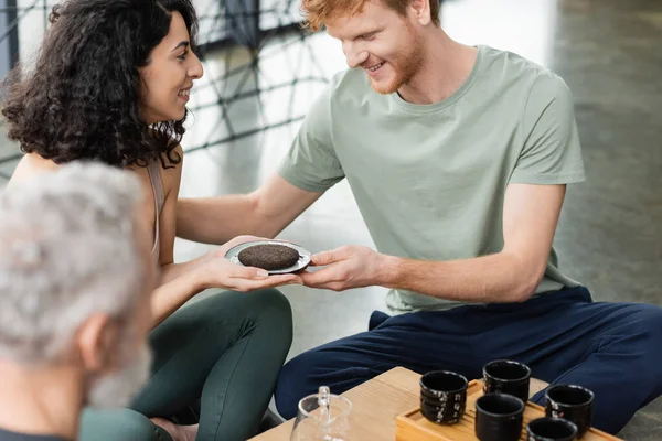 Happy middle eastern woman giving compressed puer tea to smiling redhead man in yoga studio — Stock Photo