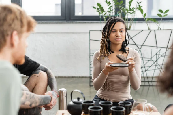 Mujer afroamericana con rastas sosteniendo té puer comprimido cerca de personas en estudio de yoga - foto de stock