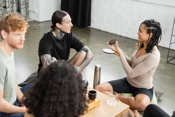 Feliz afroamericana mujer con rastas mostrando comprimido puer té a hombre tatuado en estudio de yoga - foto de stock
