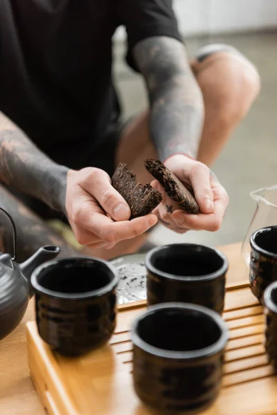 Vista recortada del hombre tatuado rompiendo el té puer comprimido cerca de la olla de té tradicional china y tazas - foto de stock