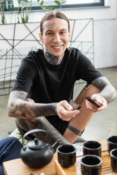 Cheerful young man with tattoos breaking compressed puer tea near traditional Chinese tea pot and cups — Stock Photo