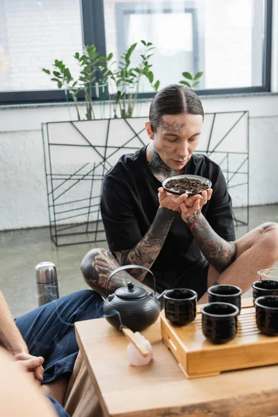 Jeune homme avec des tatouages sentant le thé puer fermenté près de brûler Palo Santo bâton et théière chinoise avec des tasses — Photo de stock
