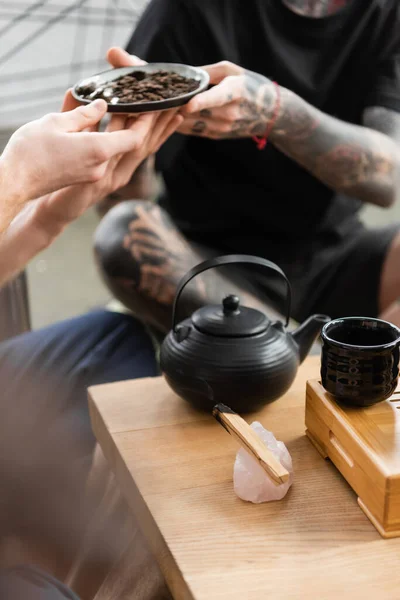 Обрезанный вид на татуированного человека, проходящего компрессионный чайник рядом с традиционным чайником и чашками в студии йоги — стоковое фото