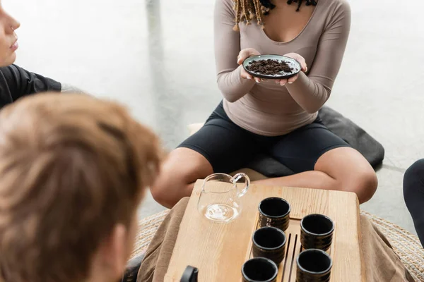 Vista ritagliata della donna afro-americana che tiene piatto con tè puro fermentato vicino agli uomini in studio di yoga — Foto stock