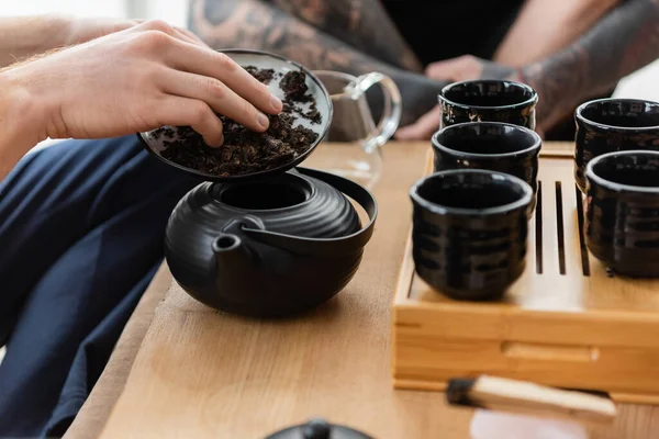 Abgeschnittene Ansicht eines Mannes, der Pu-Erh-Tee in einer traditionellen chinesischen Teekanne in der Nähe von Tassen und tätowierten Freund hinzufügt — Stockfoto