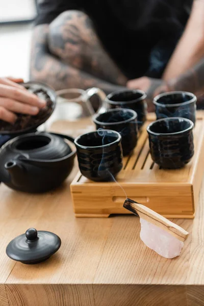 Palo Santo ardiente palo cerca del hombre añadiendo té puro en la tetera tradicional china en el estudio de yoga - foto de stock