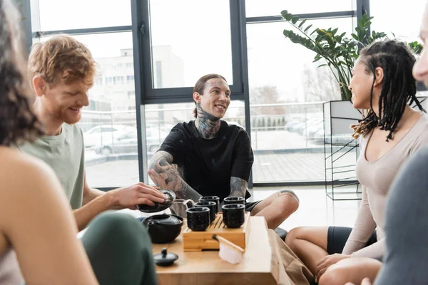 Cheerful man with tattoos sitting near interracial friends during tea ceremony in yoga studio — Stock Photo
