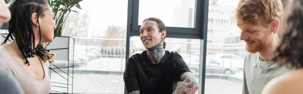 Веселий чоловік з татуюваннями, що сидить навколо міжрасових друзів в студії йоги, банер — стокове фото