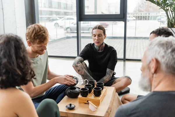Young man with tattoos sitting near interracial friends during tea ceremony in yoga studio — Stock Photo