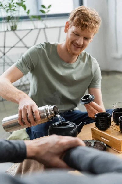 Happy redhead man holding thermos while pouring hot water and brewing puer tea in Chinese tea pot — Stock Photo
