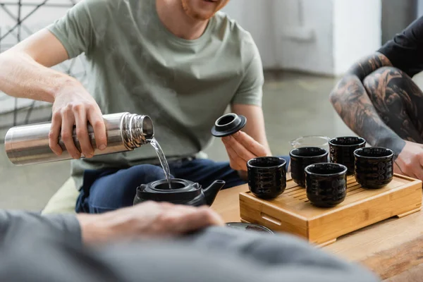 Cropped view of bearded man holding thermos while pouring hot water and brewing puer tea in Chinese tea pot — Stock Photo
