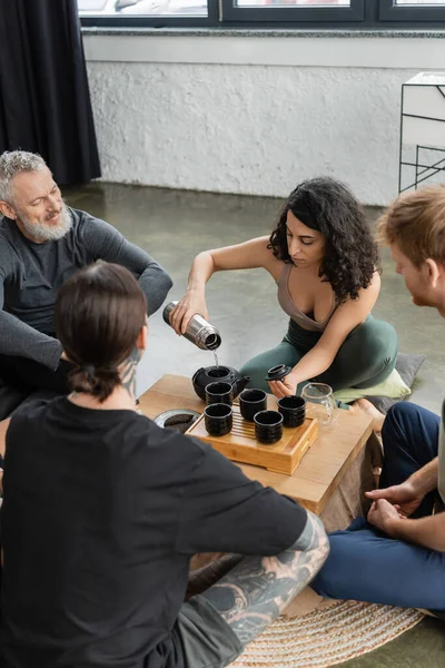 Middle eastern woman holding thermos while pouring hot water and brewing puer tea near men in yoga studio — Stock Photo