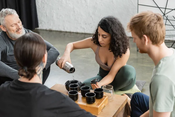 Encaracolado mulher do Oriente Médio derramando água quente de garrafa térmica e chá puer cerveja perto de homens no estúdio de ioga — Fotografia de Stock