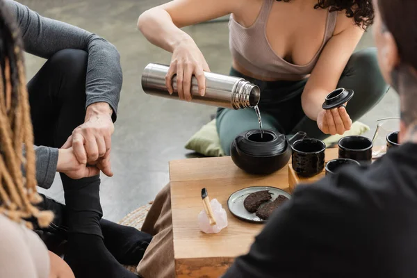 Vista cortada da mulher derramando água quente de garrafa térmica enquanto cerveja chá puer perto de pessoas no estúdio de ioga — Fotografia de Stock