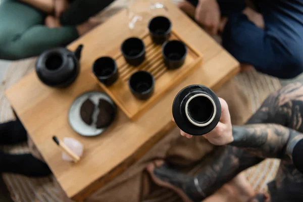 Top view of tattooed man holding cup with brewed puer tea near people in yoga studio — Stock Photo