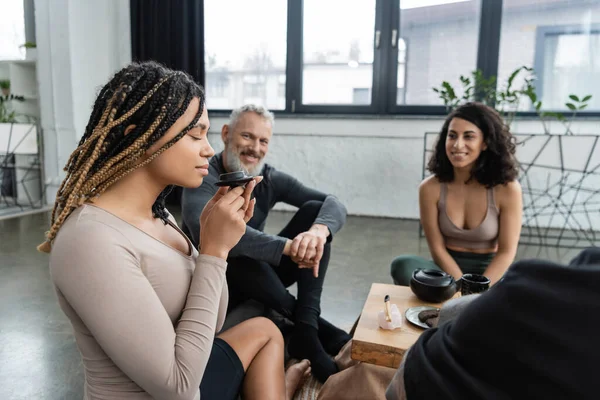 Joven afroamericana mujer con rastas disfrutando de aroma de té puro cerca de la gente sobre fondo borroso - foto de stock