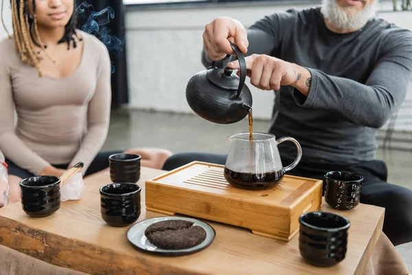 Cropped view of tattooed man pouring brewed puer tea into glass jug near african american woman on blurred background — Stock Photo